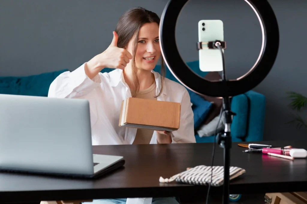 Mulher sorridente gravando vídeo com ring light e smartphone enquanto segura caixa de papelão e faz sinal de positivo