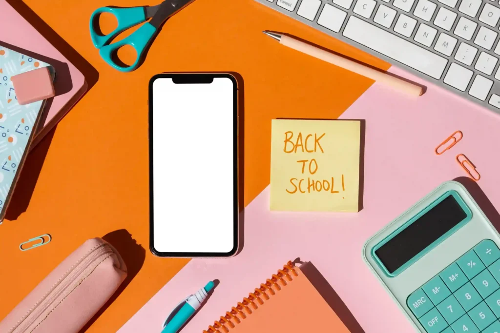 Smartphone com tela em branco rodeado por material escolar colorido, tesoura, calculadora e post-it com texto "Back to school", fundo laranja e rosa