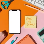 Smartphone com tela em branco rodeado por material escolar colorido, tesoura, calculadora e post-it com texto "Back to school", fundo laranja e rosa