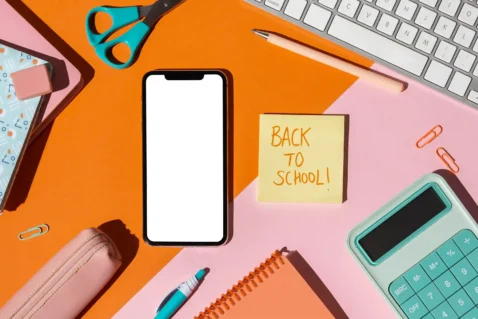 Smartphone com tela em branco rodeado por material escolar colorido, tesoura, calculadora e post-it com texto "Back to school", fundo laranja e rosa