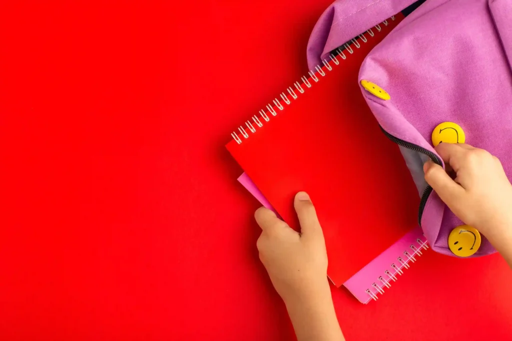 Mãos de criança retirando caderno espiral vermelho de mochila roxa com bottons amarelos, sobre fundo vermelho