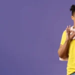 Jovem sorridente com camiseta amarela segurando mochila cinza e fazendo sinal de positivo, fundo roxo