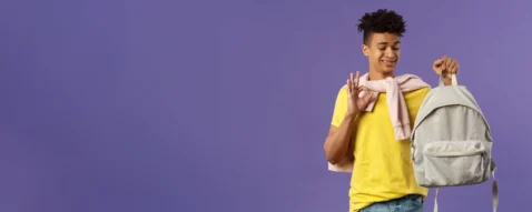 Jovem sorridente com camiseta amarela segurando mochila cinza e fazendo sinal de positivo, fundo roxo