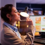 Mulher pensativa com caneta na boca diante de monitor exibindo dashboard com gráficos coloridos, iluminada por abajur em ambiente escurecido