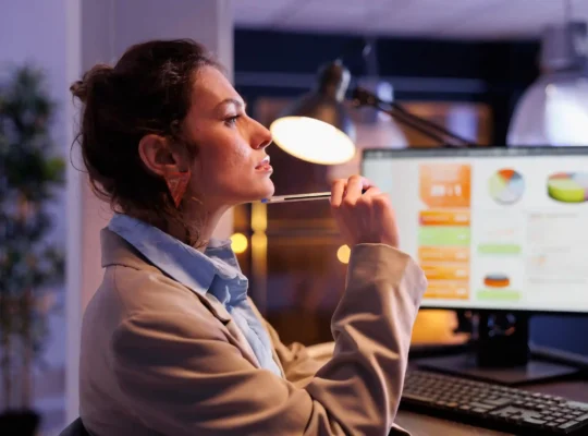 Mulher pensativa com caneta na boca diante de monitor exibindo dashboard com gráficos coloridos, iluminada por abajur em ambiente escurecido