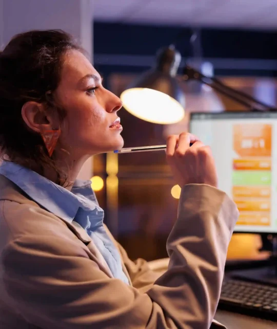 Mulher pensativa com caneta na boca diante de monitor exibindo dashboard com gráficos coloridos, iluminada por abajur em ambiente escurecido