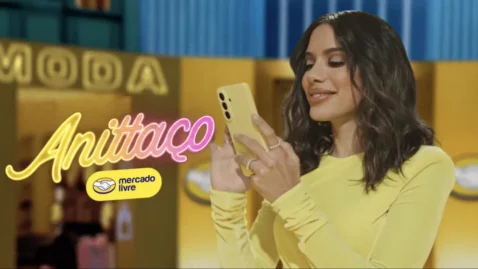 Frame de campanha publicitária Anittaço do Mercado Livre com celebridade de roupa amarela usando smartphone, letreiro "Moda" ao fundo