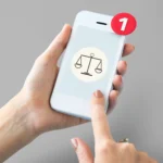Mãos segurando smartphone com ícone de balança da justiça na tela e notificação com número um, fundo cinza