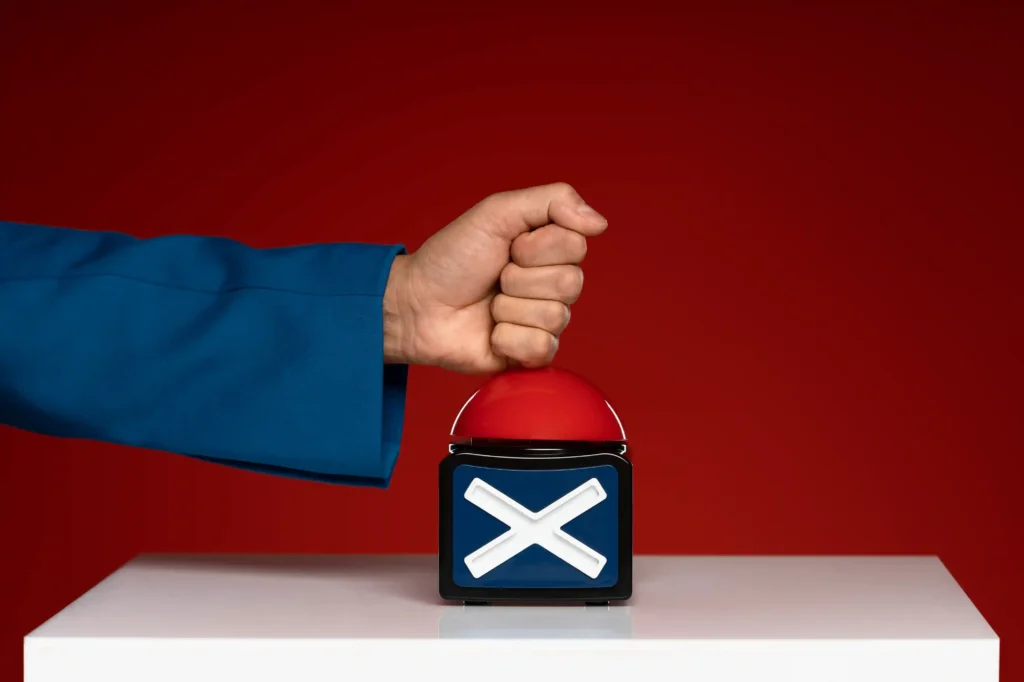 Mão com manga azul pressionando botão vermelho com símbolo de X sobre pedestal branco, fundo vermelho