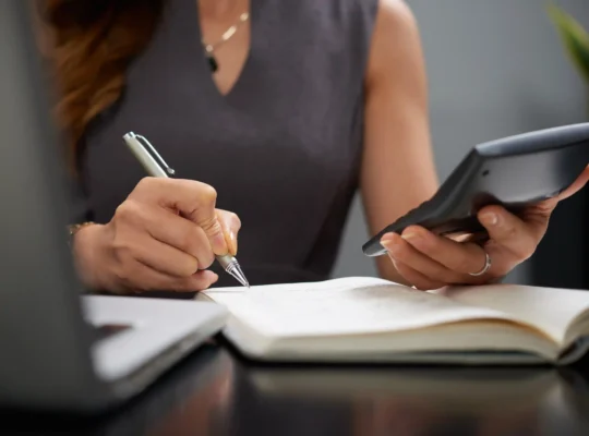 Mulher calculando números com calculadora e caderno de anotações, representando o planejamento financeiro da reforma tributária no e-commerce