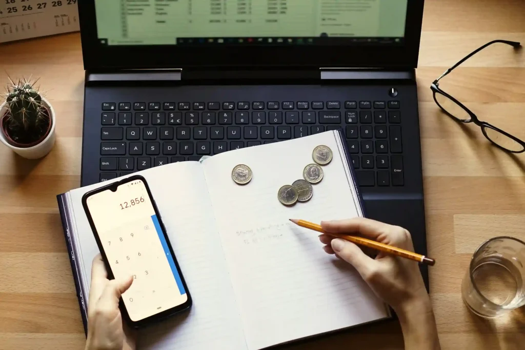 Mãos anotando em caderno com lápis e segurando celular com calculadora aberta, moedas sobre o caderno e notebook com planilha ao fundo