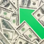 Notas de cem dólares espalhadas ao redor de uma seta verde apontando para cima, representando a alta do dólar e o aumento dos custos