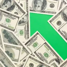 Notas de cem dólares espalhadas ao redor de uma seta verde apontando para cima, representando a alta do dólar e o aumento dos custos