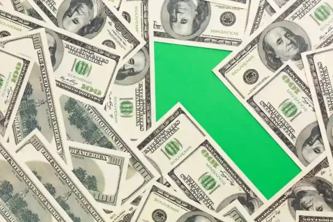 Notas de cem dólares espalhadas ao redor de uma seta verde apontando para cima, representando a alta do dólar e o aumento dos custos
