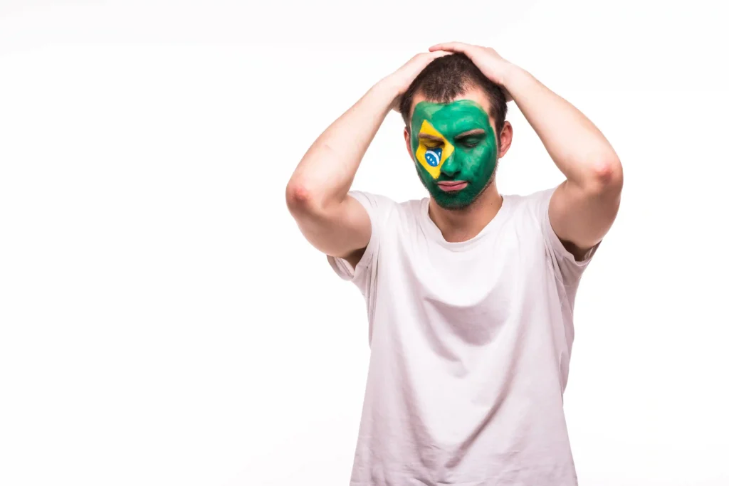 Homem com rosto pintado com as cores da bandeira do Brasil com expressão de desânimo e mãos na cabeça, fundo branco