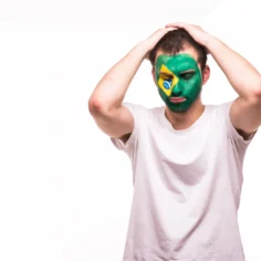 Homem com rosto pintado com as cores da bandeira do Brasil com expressão de desânimo e mãos na cabeça, fundo branco