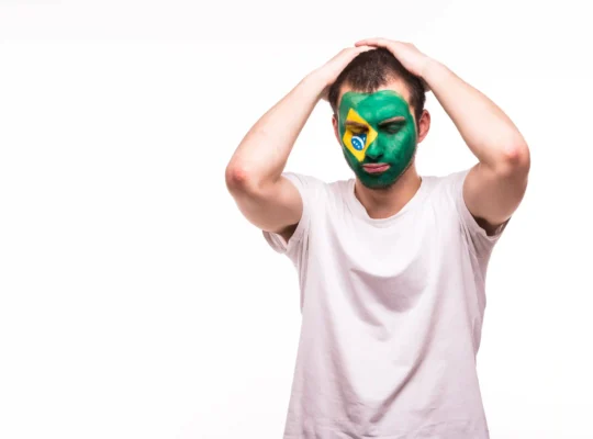 Homem com rosto pintado com as cores da bandeira do Brasil com expressão de desânimo e mãos na cabeça, fundo branco