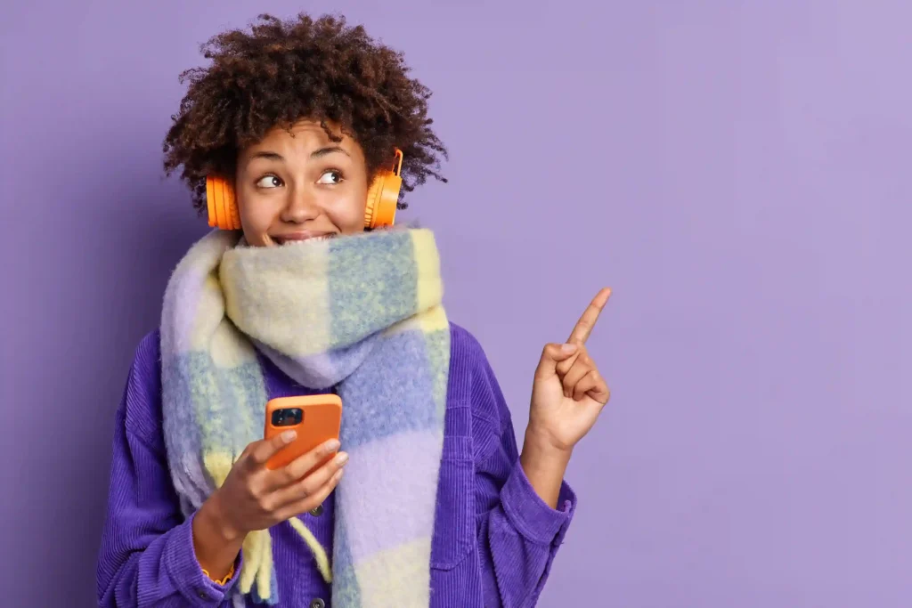 Mulher sorridente com cachecol xadrez e fone de ouvido laranja segurando smartphone e apontando para o lado, fundo roxo