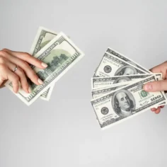 Duas mãos trocando notas de cem dólares, ilustrando o impacto do câmbio nas vendas diante das tarifas dos EUA