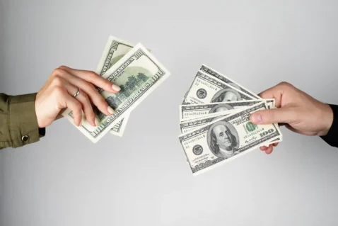 Duas mãos trocando notas de cem dólares, ilustrando o impacto do câmbio nas vendas diante das tarifas dos EUA