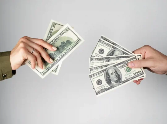 Duas mãos trocando notas de cem dólares, ilustrando o impacto do câmbio nas vendas diante das tarifas dos EUA