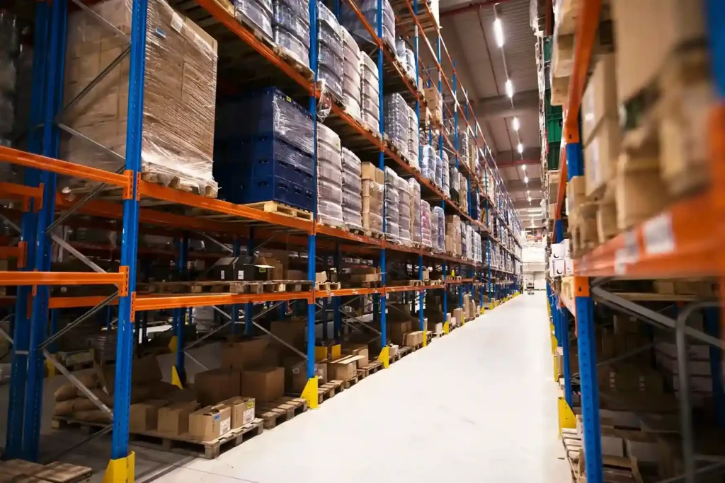 Corredor de centro de distribuição com prateleiras altas de estrutura azul e laranja repletas de paletes e caixas embaladas
