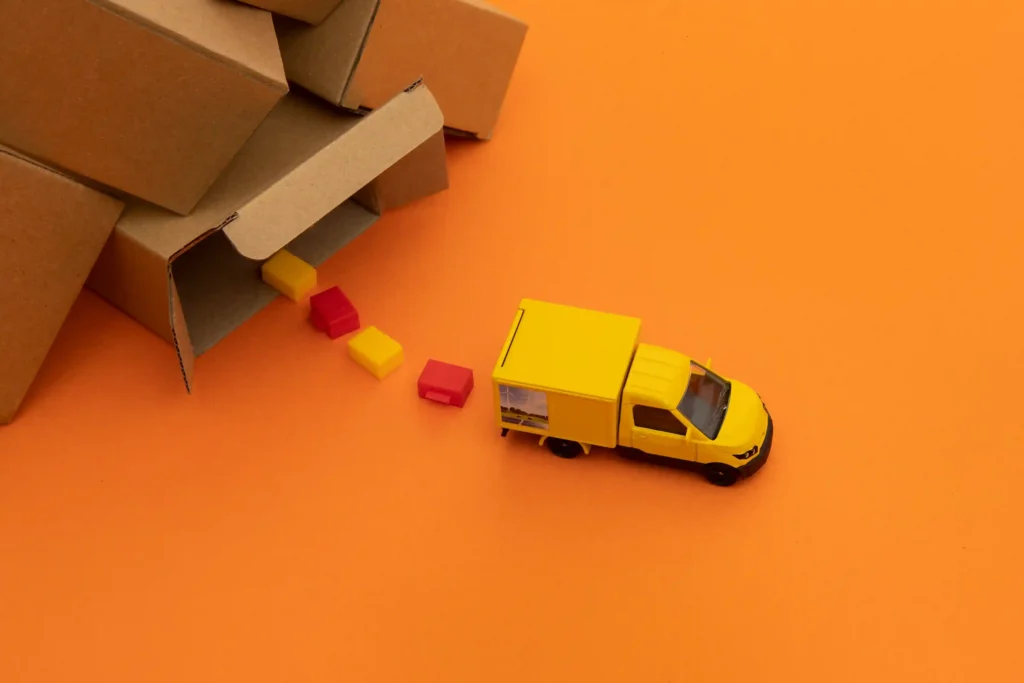 Miniatura de caminhão amarelo saindo de caixas de papelão, simbolizando a aplicação em menor escala das estratégias de Logística Mercado Livre 2026