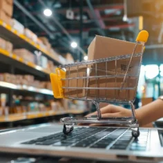 Carrinho de compras em miniatura sobre notebook em ambiente de centro de distribuição, representando a Logística Mercado Livre 2026