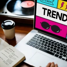 Mãos sobre notebook com tela exibindo layout colorido com a palavra "Trends" em destaque, caderno com anotações e copo de café ao lado