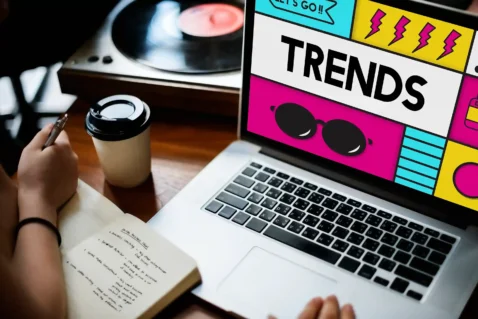Mãos sobre notebook com tela exibindo layout colorido com a palavra "Trends" em destaque, caderno com anotações e copo de café ao lado