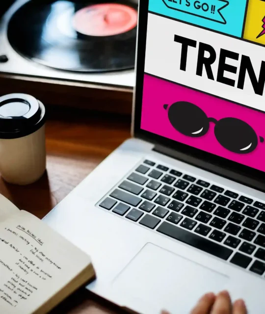 Mãos sobre notebook com tela exibindo layout colorido com a palavra "Trends" em destaque, caderno com anotações e copo de café ao lado