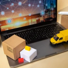 Notebook com tela exibindo mapa de rotas logísticas globais, miniaturas de caminhão amarelo, empilhadeira, avião e caixas de papelão ao redor, fundo bege