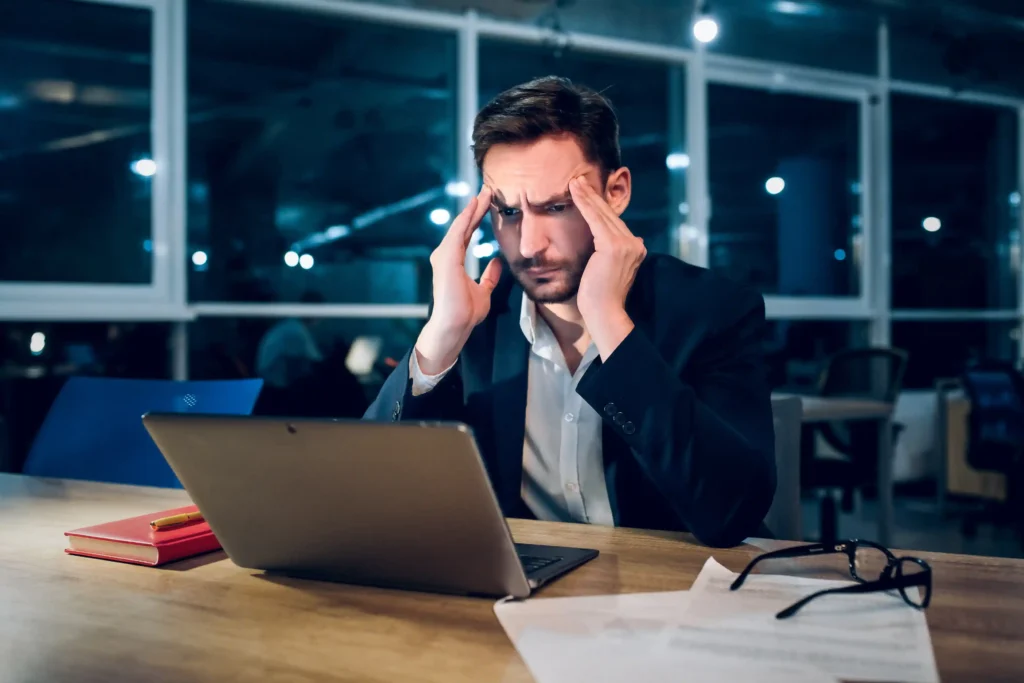 Homem de terno preto com expressão de preocupação e mãos na cabeça diante de notebook em escritório iluminado à noite