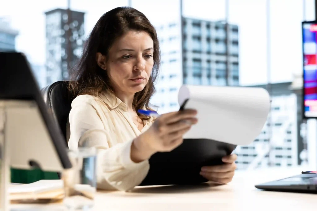 Mulher de blusa bege analisando documentos com caneta em escritório corporativo com vista para prédios ao fundo