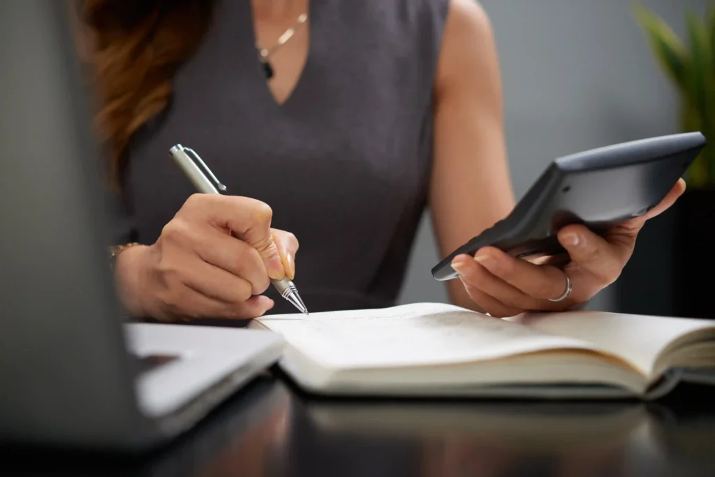 Mãos de mulher escrevendo em caderno com caneta enquanto segura calculadora, sobre mesa escura