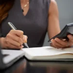 Mãos de mulher escrevendo em caderno com caneta enquanto segura calculadora, sobre mesa escura