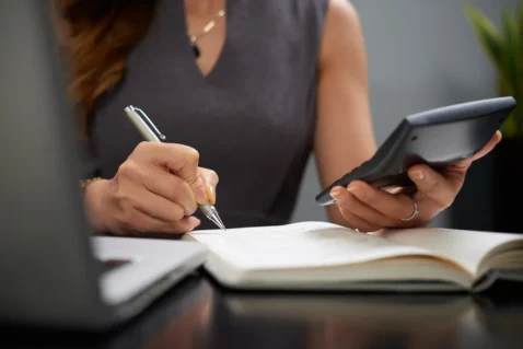 Mãos de mulher escrevendo em caderno com caneta enquanto segura calculadora, sobre mesa escura