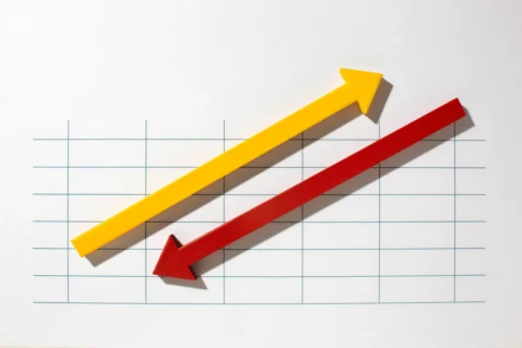 Duas setas diagonais em papel quadriculado — uma amarela apontando para cima e uma vermelha apontando para baixo — representando tendências opostas