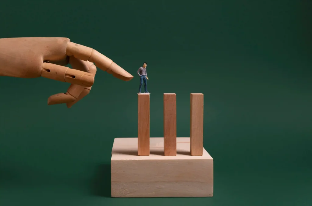 Mão de madeira aponta para miniatura de homem sobre três blocos de madeira em fundo verde