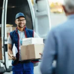 Entregador segura duas caixas diante de uma van de entregas enquanto conversa com um cliente