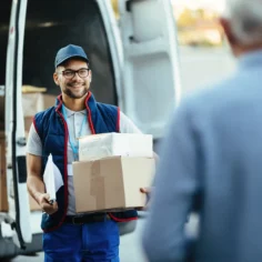 Entregador segura duas caixas diante de uma van de entregas enquanto conversa com um cliente
