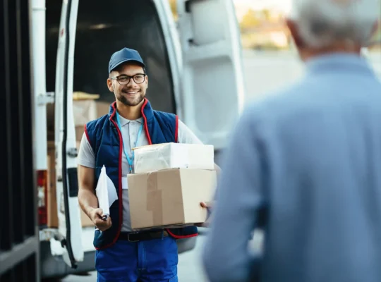 Entregador segura duas caixas diante de uma van de entregas enquanto conversa com um cliente