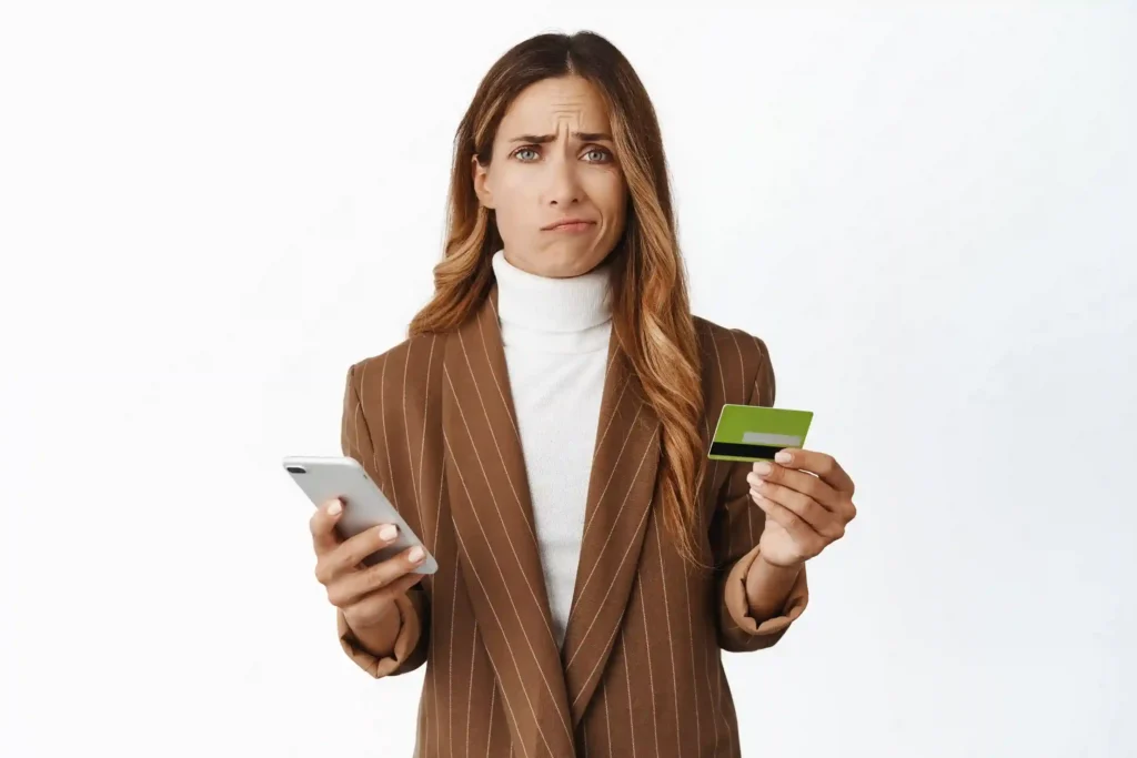 Mulher com expressão de dúvida segurando um celular e um cartão bancário