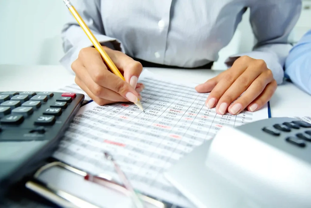 Pessoa usa lápis para conferir uma planilha impressa ao lado de duas calculadoras