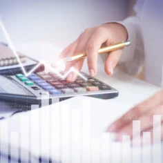 Pessoa usa calculadora sobre uma mesa enquanto gráficos de linhas e barras aparecem sobrepostos à imagem