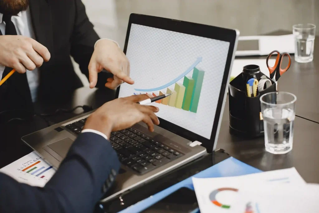 Pessoas apontam para um gráfico de barras em crescimento exibido na tela de um notebook sobre uma mesa
