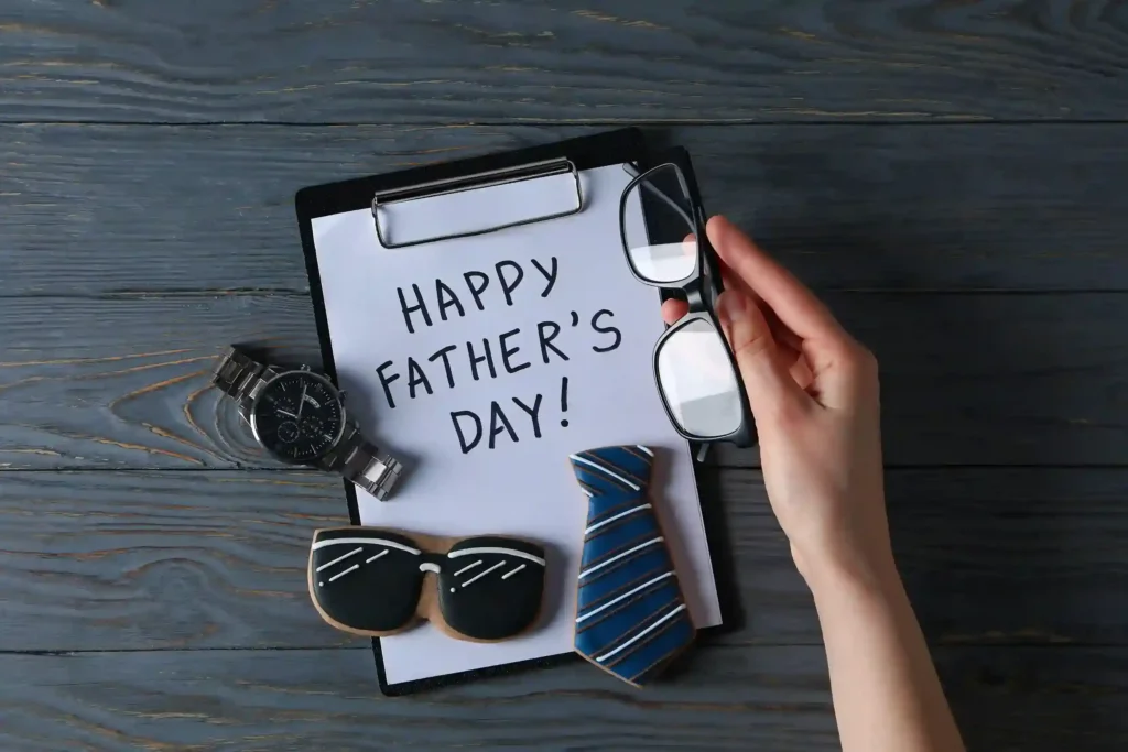 Prancheta com a mensagem “Happy Father’s Day”, relógio, gravata e biscoito em formato de óculos sobre uma mesa de madeira, enquanto uma mão segura óculos de grau