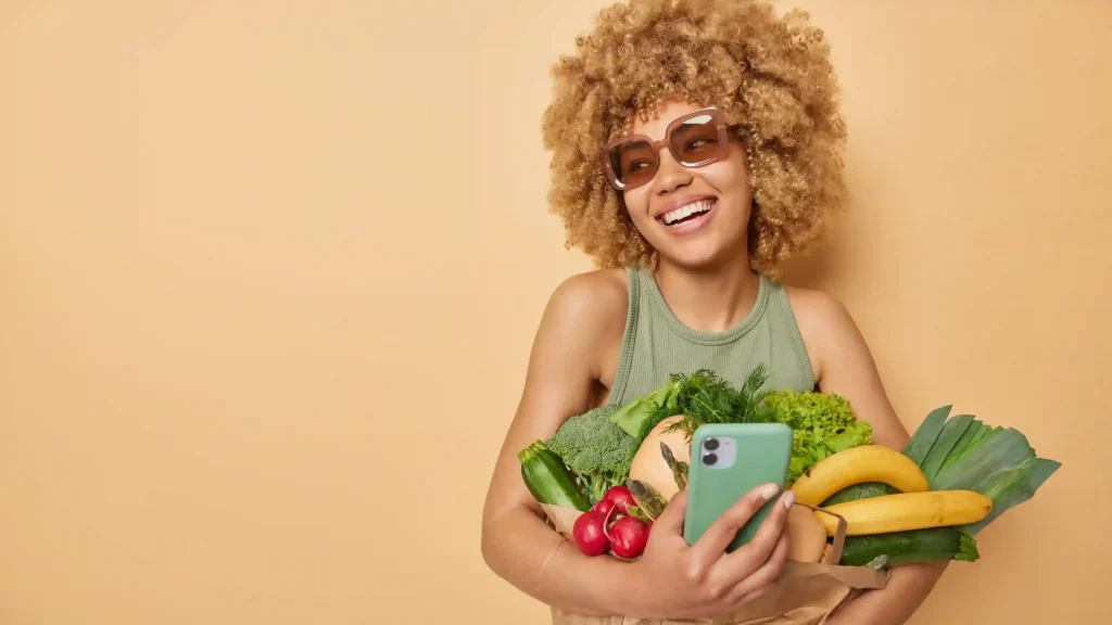 Mulher segurando frutas e vegetais enquanto usa o celular
