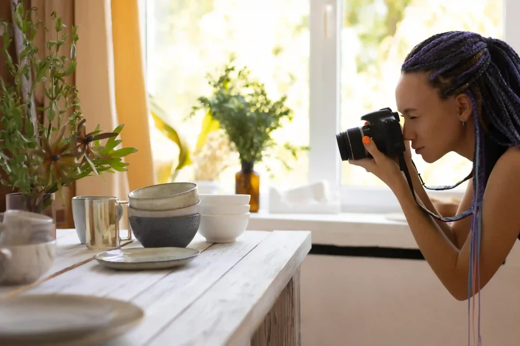 Fotógrafa usando uma câmera para fotografar louças, copos e outros objetos organizados sobre uma mesa, representando a produção de fotos de produtos para vendas no e-commerce