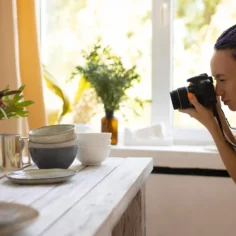 Fotógrafa usando uma câmera para fotografar louças, copos e outros objetos organizados sobre uma mesa, representando a produção de fotos de produtos para vendas no e-commerce